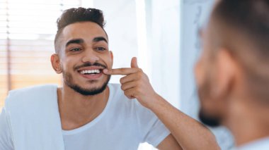 Genç bir adam parlak bir banyoda aynanın önünde duruyor, gülümsüyor ve dişlerini işaret ediyor. Basit bir beyaz gömlek giyiyor ve diş bakımından memnun görünüyor..