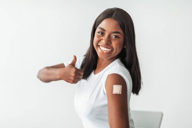 Covid 'e karşı etkili bir aşı. Neşeli, heyecanlı, gülümseyen, aşılanmış kollarını omzuna yapıştırarak gösteren siyah kadın hastanın portresi. Vurulduktan ve başparmak hareketiyle, Beyaz Stüdyo