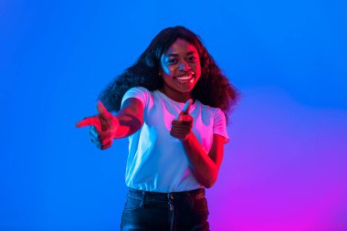 Genç bir kadın stüdyoda poz verirken sevinçle gülümsüyor. Beyaz gömlek ve siyah pantolon giyiyor, enerjik bir kişilik sergiliyor canlı bir mavi ve pembe arka plana karşı.