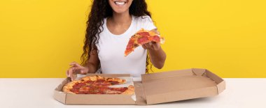 Bir insan masada otururken bir dilim pizza tutmaktan mutlu olur. Arka plan parlak sarı, neşeli bir atmosfer yaratıyor. Pizza kutusu açık, leziz dilimleri ortaya çıkarıyor..