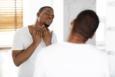 Bir adam iyi aydınlatılmış bir banyoda duruyor, tımar etmeye odaklanıyor. Aynadan yansımasına bakarken nazikçe çene hattına dokunur ve ilerideki güne hazırlanır..