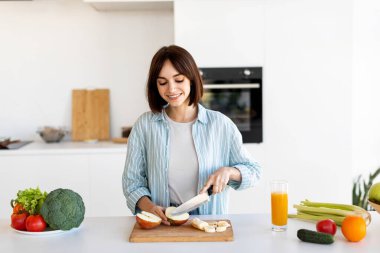 Gülümseyen bir kadın taze meyve ve sebzelerle dolu modern bir mutfakta elma dilimliyor. Sağlıklı atıştırmalıklar hazırlarken ferahlatıcı bir portakal suyu içiyor..