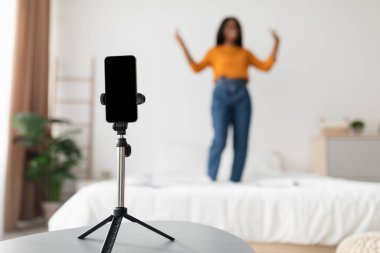 Parlak bir odada, bir kadın yatakta duruyor, konuşma yaparken el kol hareketi yapıyor. Tripodun üzerindeki bir akıllı telefon, hafif bir arkaplanda sunumunu yakalar..