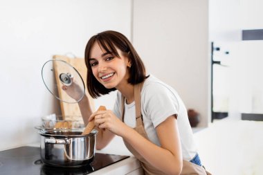 Neşeli bir suratı olan genç bir kadın modern bir mutfakta ocakta demlik karıştırıyor. Doğal ışık odayı dolduruyor, yemek pişirme zevkini vurguluyor..