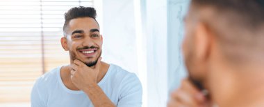 Bir adam aynada yansımasına bakıyor, çenesine dokunurken özenle gülümsüyor. Perdelerin arasından gelen güneş ışığıyla ortam aydınlık, neşeli bir atmosfer yaratıyor..