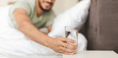 Bir adam bir komodinin üzerine yerleştirilmiş bir bardak suya doğru elini uzatıyor. Konforlu yatak odasında yumuşak yatak örtüsü var. Tazelenmiş ve memnun görünüyor..