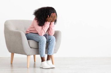 Kıvırcık saçlı genç bir kız açık gri bir sandalyede oturuyor, elleriyle yüzünü örtüyor. Oda parlak ve basit, kasvetli bir atmosfer yaratıyor..