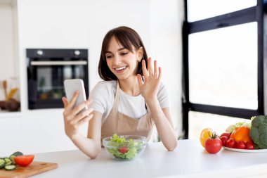 Neşeli genç bir kadın mutfak masasında otururken telefonuna el sallıyor. Elinde canlı sebzelerle çevrili taze salata malzemeleri var..