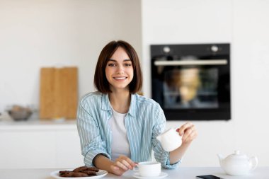 Bir kadın mutfak tezgahında oturmuş, fincana çay dökerken gülümsüyor. Yanında bir tabak kurabiye var ve mutfak temiz ve modern bir tasarıma sahip. Sıcak öğleden sonra ışığı boşluğu dolduruyor..