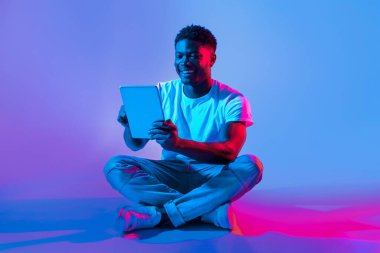 Genç bir adam tabletle etkileşime girerken yerde bağdaş kurup gülümsüyor. Canlı mavi ve pembe ışıklar canlı bir atmosfer yaratarak dijital deneyimden zevk almasını sağlıyor..