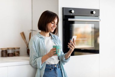 Parlak bir mutfakta, kısa saçlı genç bir kadın bir deniz mavisi bardağı tutuyor, akıllı telefonuna bakarken gülümsüyor. Gündüz giyinmiş ve güneş ışığı gibi rahatlamış..