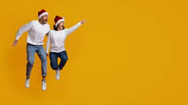 Noel Baba şapkası takıp şenlik sezonunu kutlayan bir çift sevinçle zıplıyor. Neşeli pozları ve parlak elbiseleriyle heyecan ve mutluluğu cesur sarı ayarıyla ifade ederler..