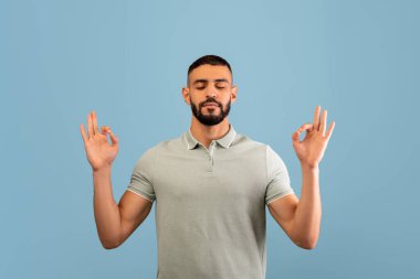 Genç bir adam gözleri kapalı, huzurlu bir şekilde oturur, farkındalık meditasyonu yapar. Elleri sakin bir jest yapıyor, açık mavi bir arkaplana karşı sükunet yayıyor..