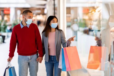 Bir çift, vitrinlere bakarken renkli alışveriş torbaları taşıyarak el ele yürür. Yüz maskesi takıyorlar ve alışveriş merkezlerinde alışveriş yapmaktan zevk alıyor gibi görünüyorlar..