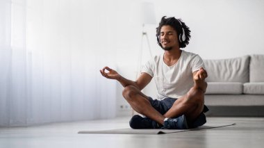 Rahatlamış bir siyah adam evdeki yoga minderinde nilüfer pozisyonunda oturuyor, kablosuz kulaklık takıyor. Gözleri kapalı gülümser, meditasyon sırasında sükunet ve huzuru somutlaştırır..