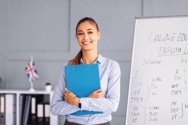 Bir bayan öğretmen mavi bir klasörle kameraya gülümsüyor. İngilizce içeriğini gösteren bir yazı tahtasıyla çevrimiçi eğitim ortamında ve arka planda bir bayrak var..