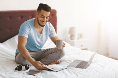 Gülen adam elinde kahve fincanı tutarken bacak bacak üstüne atarak oturuyor. Parlak, rahat bir yatak odasında dizüstü bilgisayarının yanındaki deftere bakar ve sabah rutininin tadını çıkarır..