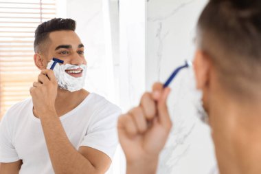 Modern bir banyoda, bir adam jiletle tıraş oluyor, yansımasına gülümsüyor. Tıraş kremi sürüyor ve bakım rutini sırasında mutlu görünüyor. Doğal ışık boşluğu doldurur..