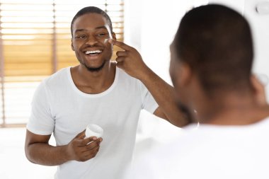 Bir adam aynanın karşısında duruyor, yüzüne cilt ürünleri sürerken gülümsüyor. Parlak banyoda ahşap panjurlar ve temiz ve modern bir tasarım var..