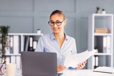 Gözlüklü genç bir kadın masasında belgeleri incelerken gülümsüyor. Bitkiler ve raflarla dolu parlak, çağdaş bir ofiste dizüstü bilgisayarına odaklanmış durumda..