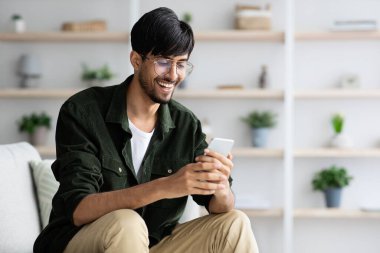 Oturma odasında rahat bir şekilde oturan bir adam akıllı telefonunu kullanırken gülümsüyor. Mekan parlak ve bitki ve raflarla süslenmiş. Hoş bir atmosfer yaratıyor..