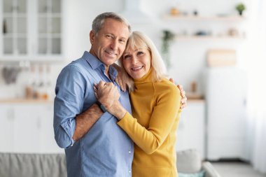 Mutlu çift oturma odalarında durur, gülümsemeyle sevgi dolu bir kucaklaşmayı paylaşırlar. Parlak güneş ışığı pencerelerden süzülerek sıcak bir atmosfer yaratır..