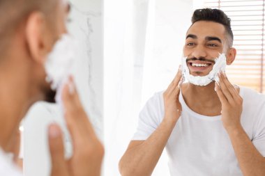 Bir adam aynanın karşısında duruyor, sakalına tıraş kremi sürerken gülümsüyor. Banyo doğal ışıkla aydınlanmış, tımar için yeni bir atmosfer yaratıyor..