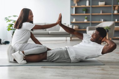 Mutlu gülen genç siyah çift evde karın kasları üzerinde çalışıyor, mekik çeken adam, erkek bacakları üzerinde oturan kadın oturma odasında beşlik çakıyor. Spor, ev spor ve destek konsepti