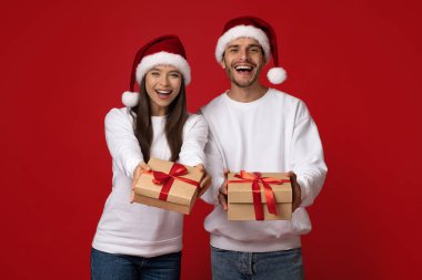 Noel Baba şapkası takan neşeli bir çift canlı kırmızı bir zemin üzerinde duruyor, her biri süslenmiş bir hediye kutusu tutuyor, bayram neşesi ve heyecanı yayıyorlar..