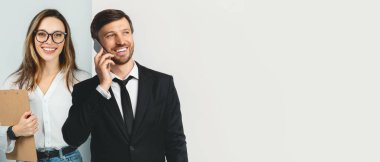 İki profesyonel bir iş görüşmesi yapıyor. Bir kişi telefonda konuşurken gülümsüyor, diğeri de bir not panosu tutuyor. Parlak bir ortamda görevlerine odaklanmış görünüyorlar..