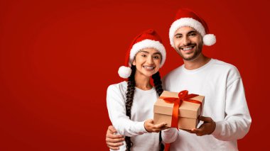 Mutlu bir çift birlikte duruyor, ikisi de Noel Baba şapkası takıyor, ellerinde kırmızı kurdeleli bir hediye var. Canlı kırmızı arka plan, Noel 'i kutlarken neşeli ifadelerini artırıyor..