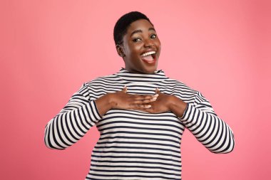 Neşeli bir kadın ellerini göğsüne koyup, gülümseyerek ayakta duruyor. Çizgili bir süveter giyiyor, ve parlak pembe arka plan onun canlı görünümüne ekleniyor..