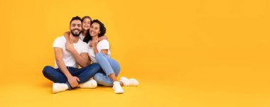 Bir aile sarı bir zeminde birlikte oturur, kucaklaşır ve gülümser. Neşeli bir anı paylaşıyorlar, aşkı ve birlikteliği yansıtıyorlar..