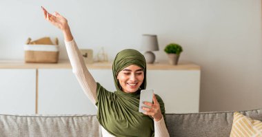 Yeşil başörtüsü takan bir kadın oturma odasından bir video görüşmesi yaparken gülümsüyor. Tebrik etmek için elini kaldırır, konuşma sırasında heyecanını gösterir..