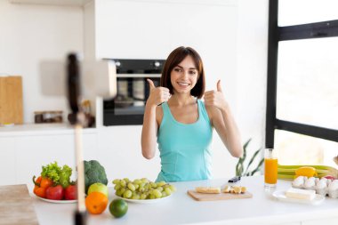 Güneşli bir mutfakta, genç bir kadın tezgahın üzerinde renkli meyve ve sebzelerle çevrili bir şekilde başparmağıyla poz verir ve sağlıklı bir yemek pişirme anını gösterir..
