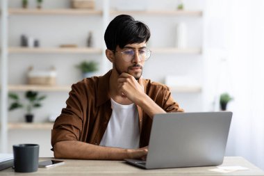 Kahverengi gömlekli bir adam ahşap bir masada oturuyor, dizüstü bilgisayarına odaklanmış. Güneş ışığı pencereden süzülerek, arka planda bitkiler olan iyi organize edilmiş bir çalışma alanını aydınlatıyor.