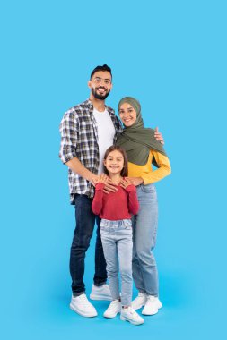 Neşeli bir aile, parlak mavi bir zemine gülümsüyor. Baba işaretli bir gömlek giyer, anne tesettürlü ve renkli bir kıyafet giyer, ve kızları kırmızı kazak giyer..