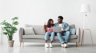 Çevrimiçi ödeme. Oturma odasındaki koltukta oturan siyah eşleri internetten alışveriş yapıyor, bilgisayar ve elektronik bankacılık kullanan Afrikalı genç çiftler, kopyalama alanı kullanıyor.