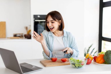 Bir kadın video bağlantısı kurarken taze bir salata hazırlamak için domatesleri zevkle dilimliyor. Modern mutfak canlı sebzeler ve ışıkla dolu..