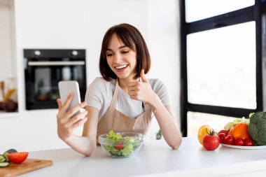 Bir kadın mutfak tezgahında bir kase taze salatayla oturuyor. Seve seve selfie çeker, sağlıklı yemeğinin tadını çıkarır. Yakında parlak sebzeler görülebilir..