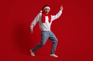 Noel Baba şapkası ve kırmızı eşarp takan bir adam parlak kırmızı bir arka planda neşeyle dans ediyor. Şenlik kıyafetleri Noel ruhunu yansıtıyor, bayram sezonunu kutluyor..