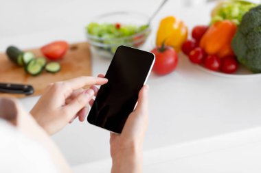 Parlak bir mutfakta bir insan renkli sebze salatası hazırlarken akıllı telefonuna bakar. Masada salatalık, domates ve dolma biber gibi taze malzemeler var..