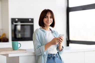 Parlak bir mutfakta duran genç bir kadın akıllı telefonu tutarken gülümsüyor. Sıradan bir kıyafet giyer ve tezgaha yaslanır. Evdeki zamanının tadını çıkarır..