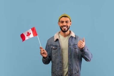 Kot ceket ve yeşil bereli bir adam mavi bir fonda duruyor. Elinde küçük bir Kanada bayrağı tutuyor ve neşe ve gurur ifade eden baş parmağını kaldırıyor..