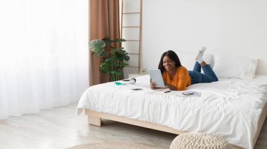 Bir kadın tablet kullanırken gülümseyerek yatağın üzerinde yüzüstü yatıyor. Odada bitkiler, parlak güneş ışığı ve rahatlatıcı bir dekor var. Rahat bir atmosfer yaratıyorlar..