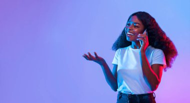 Kıvırcık saçlı neşeli bir kadın bir telefon görüşmesi yapıyor. Arka planda parlak, renkli bir ışık var, canlı bir atmosfer yaratıyor, neşeli tavırlarını geliştiriyor..