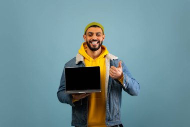 Kot ceketli ve sarı kapüşonlu bir adam mavi arka planda duruyor. Geniş bir gülümseme sergiliyor ve elinde bir dizüstü bilgisayarla olumlu bir tavır sergiliyor..