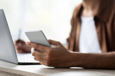 Bir insan ahşap bir masada dizüstü bilgisayar üzerinde çalışırken bir elinde akıllı telefon tutuyor. Olay yeri aydınlık bir alanda geçiyor. Bu da sakin bir çalışma ortamı olduğunu gösteriyor..