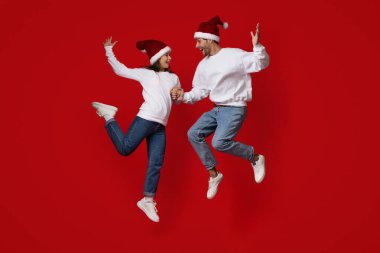 Noel süveteri ve Noel Baba şapkaları giyen neşeli bir çift havaya zıplıyor, el ele tutuşuyorlar, canlı kırmızı bir arka planda, bayram ruhunu kutluyorlar..