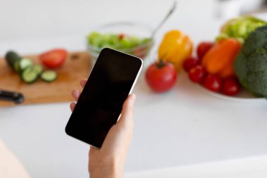 Bir insan bir elinde akıllı telefon tutarak domates, salatalık ve dolma biber gibi taze sebzelerle dolu bir mutfakta yemek hazırlar..